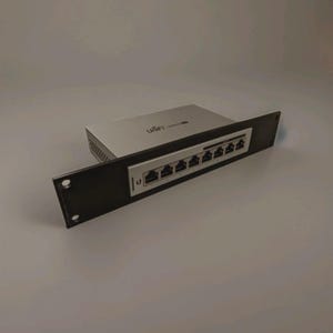 Könnte beinhalten: Ein silberner und schwarzer Netzwerk-Switch mit dem Wort "UniFi" auf der Oberseite. Der Switch verfügt über mehrere Ethernet-Anschlüsse und ist auf einer schwarzen Rackblende montiert. Die Blende hat zwei Befestigungslöcher.