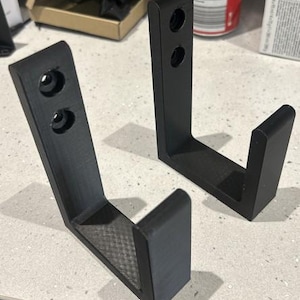 May include: Two black metal L-shaped brackets. Each bracket has two holes near the top and a hook shape at the bottom. The brackets are designed for mounting and support.