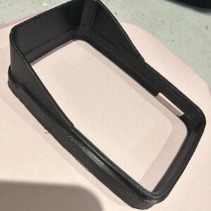 May include: A black, rectangular 3D-printed object with a raised, angled top and a hollow interior. The object has a textured surface and appears to be a frame or housing for another component. The item is sitting on a light pink surface.