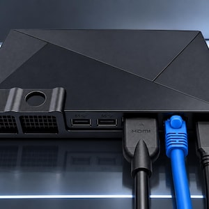 Op de afbeelding: Een zwart, rechthoekig media-streamingapparaat met een geometrisch bovenoppervlak. Het voorpaneel is voorzien van twee USB-poorten, een ventilatierooster en drie aangesloten kabels: een zwarte HDMI-kabel, een felblauwe Ethernet-kabel en een zwarte stroomkabel.