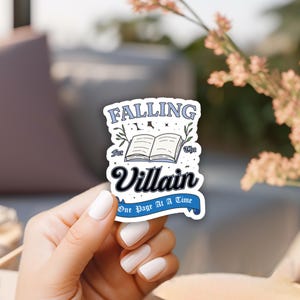 May include: A white sticker with blue and black text that reads "Falling for the Villain One Page At A Time". The sticker features a cartoon image of an open book with stars and vines.