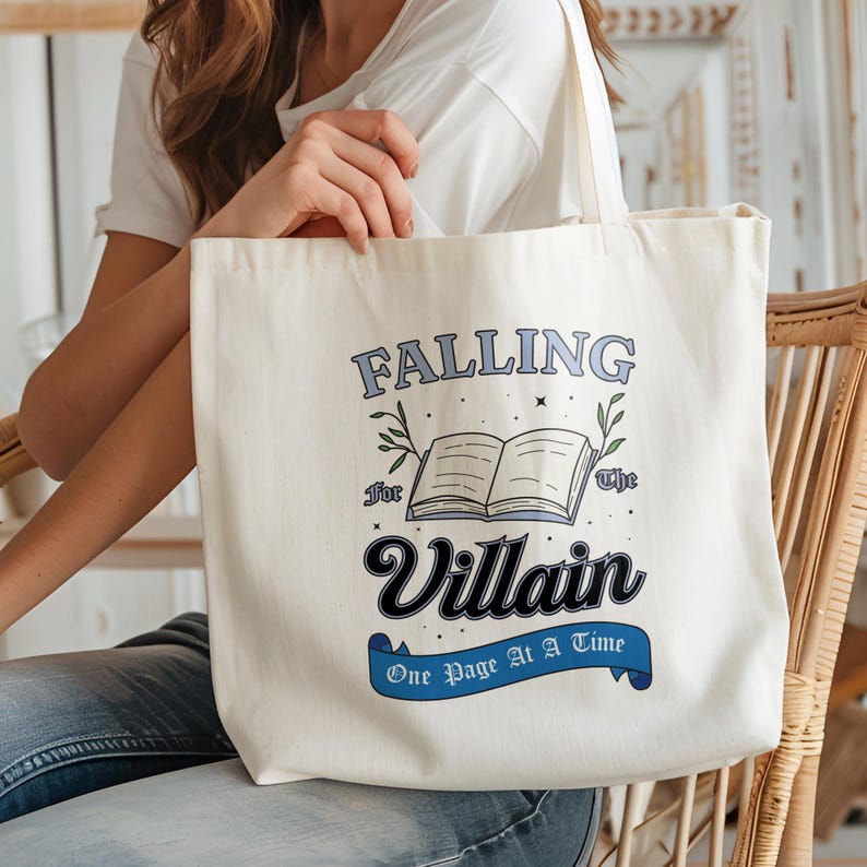 May include: A white tote bag with a blue and black design that says "Falling for the Villain One Page at a Time". The design includes a book with a blue cover and green leaves.