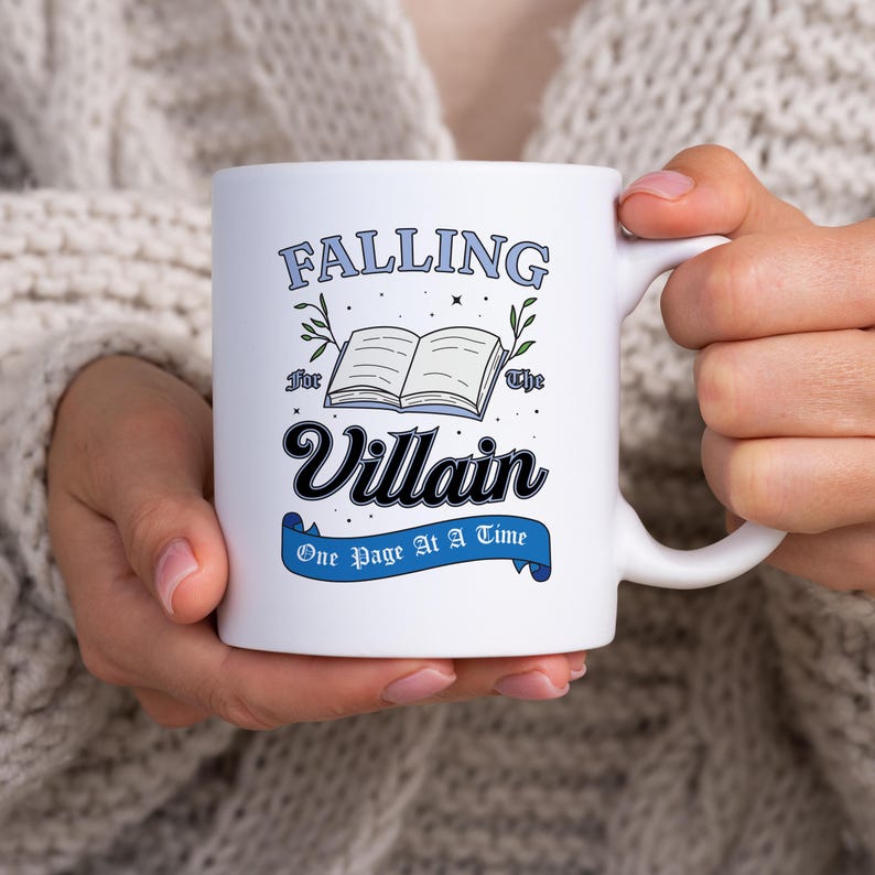 May include: White ceramic mug with a blue and black design. The mug features the text "Falling for the Villain One Page at a Time" with an illustration of an open book.