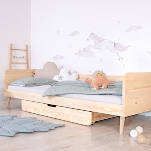 Op de afbeelding: Een licht houten kinderbed met een witte eenhoorn en wolk wanddecoratie. Het bed heeft een uittrekbare lade en een lichtgroen tapijt op de vloer.