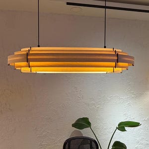 May include: A modern, layered wooden pendant light fixture. The light has a rectangular shape with multiple horizontal wooden slats, creating a tiered effect. The fixture hangs from black cords, and the light source is warm.