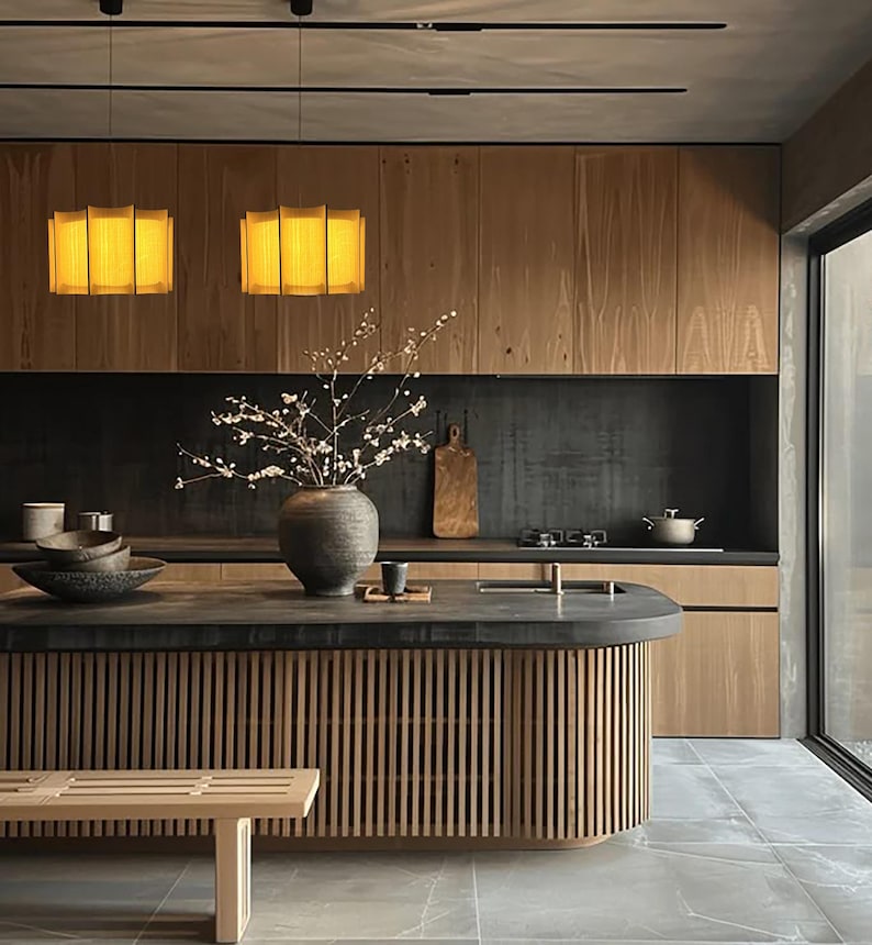 May include: A modern kitchen with wooden cabinetry and a central island. The island features a slatted wood design and a dark countertop. Two pendant lights hang above, and a bench sits in front of the island. A large vase holds branches.