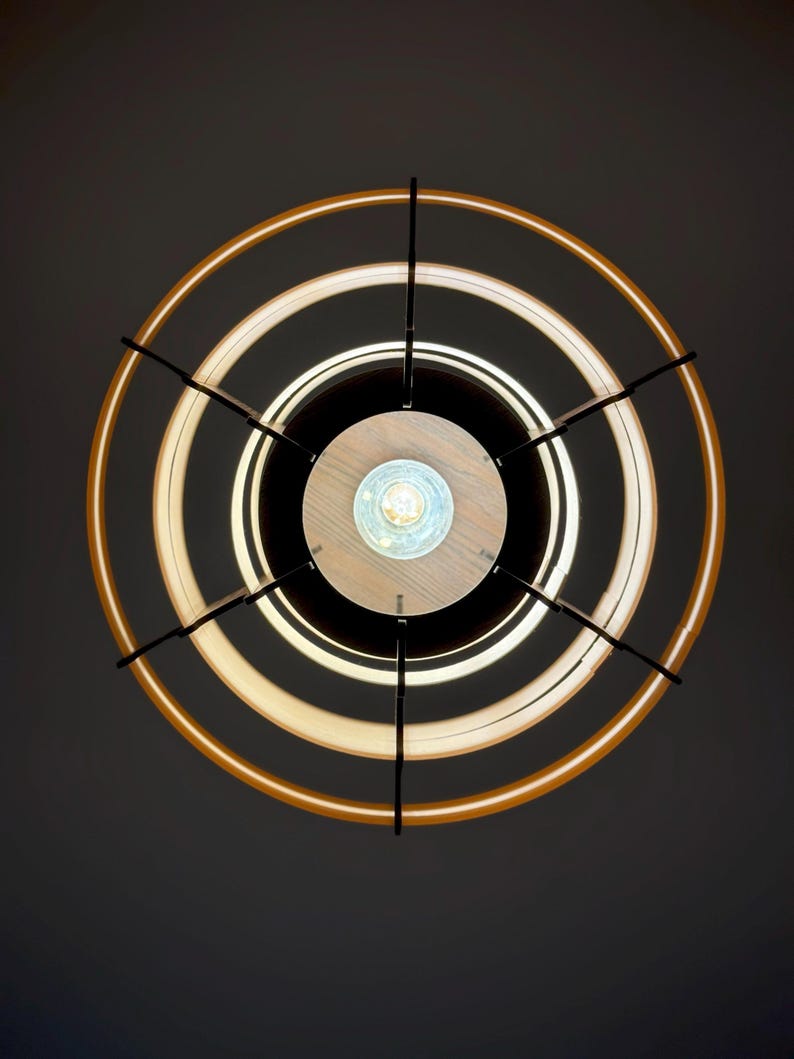 Puede incluir: Una moderna l&aacute;mpara de techo circular, vista desde abajo. Presenta un centro de madera, rodeado de anillos negros, blancos y &aacute;mbar iluminados. Varillas met&aacute;licas negras se extienden desde el centro, conectando los anillos. Dise&ntilde;o contempor&aacute;neo.