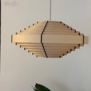 May include: A light wood pendant lamp with a layered, geometric design. The lamp has a series of horizontal wooden slats, creating a spiral effect. Black accents are visible between the wooden layers. The word "studio" is faintly visible in the background.