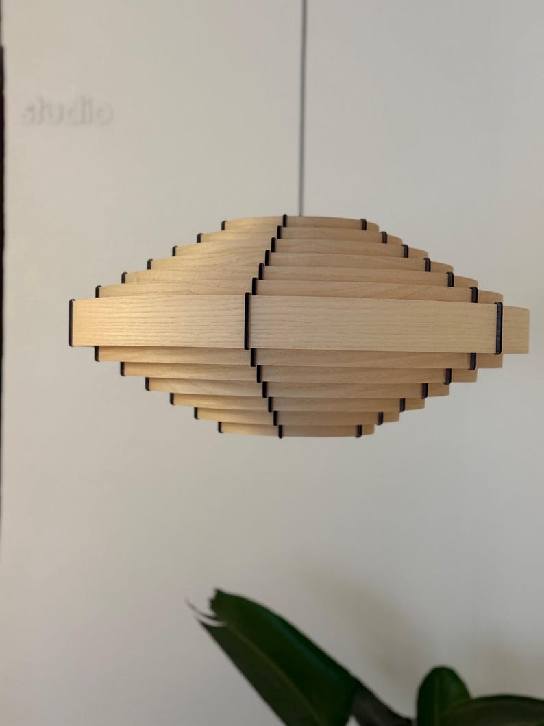 May include: A light wood pendant lamp with a layered, geometric design. The lamp has a series of horizontal wooden slats, creating a spiral effect. Black accents are visible between the wooden layers. The word "studio" is faintly visible in the background.