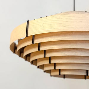 May include: A modern, layered wooden pendant light fixture. The light fixture features multiple curved, light-colored wooden slats held together by dark metal supports. The design is a spiral shape, with a black cord suspending the light.
