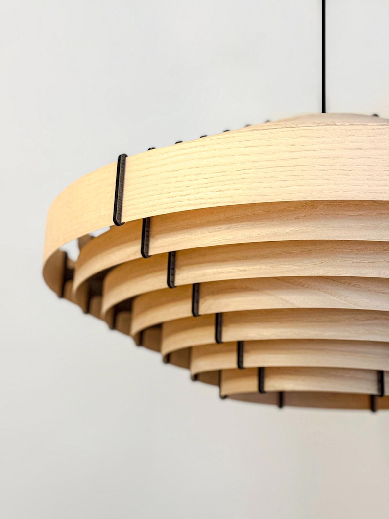 May include: A modern, layered wooden pendant light fixture. The light fixture features multiple curved, light-colored wooden slats held together by dark metal supports. The design is a spiral shape, with a black cord suspending the light.