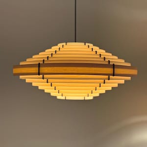 Könnte beinhalten: Eine moderne, geometrische Pendelleuchte. Die Leuchte besteht aus geschichtetem, hellfarbigem Holz mit einem dunkleren Holzband in der Mitte. Die Leuchte hängt an einem schwarzen Kabel, und das Licht ist eingeschaltet und beleuchtet das geschichtete Design.