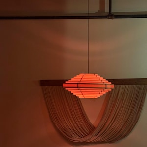 May include: A modern, geometric pendant light fixture with a warm orange glow. The light is suspended from the ceiling and is made of layered wooden slats. Below the light is a draped, woven wall hanging.