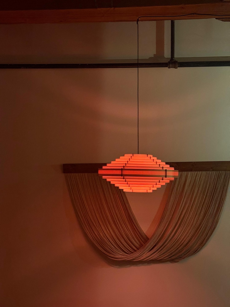 May include: A modern, geometric pendant light fixture with a warm orange glow. The light is suspended from the ceiling and is made of layered wooden slats. Below the light is a draped, woven wall hanging.