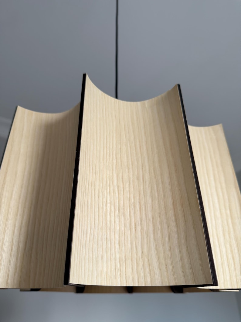 May include: A modern pendant light fixture with a unique design. The light features curved wooden panels with a light wood grain finish and black edges. The fixture hangs from a black cord, showcasing a contemporary aesthetic.