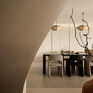 Puede incluir: Comedor moderno con una mesa rectangular negra y varias sillas. Dos l&aacute;mparas colgantes cuelgan sobre la mesa. La habitaci&oacute;n tiene una paleta de colores neutros con una pared curva en primer plano y ramas decorativas.