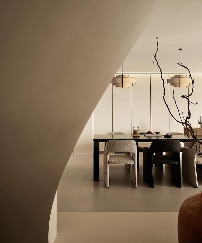 Puede incluir: Comedor moderno con una mesa rectangular negra y varias sillas. Dos l&aacute;mparas colgantes cuelgan sobre la mesa. La habitaci&oacute;n tiene una paleta de colores neutros con una pared curva en primer plano y ramas decorativas.