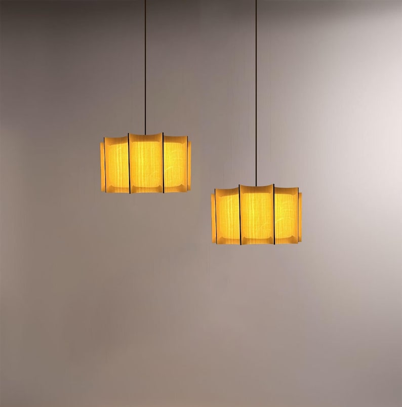 May include: Two rectangular pendant lights with a warm yellow glow. Each light has a wooden frame with vertical black lines and a fabric shade. The lights hang from black cords against a neutral background.