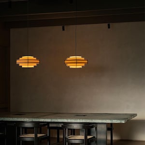 Puede incluir: Dos l&aacute;mparas colgantes con un dise&ntilde;o geom&eacute;trico en capas. Las luces tienen un brillo amarillo c&aacute;lido que emana del interior de los marcos de madera. Est&aacute;n suspendidas sobre una mesa larga y oscura con sillas a juego.