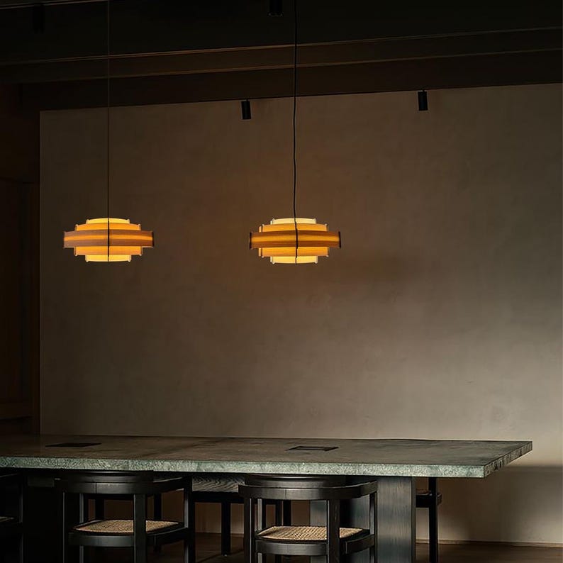 Puede incluir: Dos l&aacute;mparas colgantes con un dise&ntilde;o geom&eacute;trico en capas. Las luces tienen un brillo amarillo c&aacute;lido que emana del interior de los marcos de madera. Est&aacute;n suspendidas sobre una mesa larga y oscura con sillas a juego.