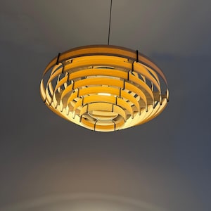 May include: A modern, wooden pendant light fixture. The light has a layered, circular design with warm, yellow light emanating from within. The fixture is suspended from a black cord and mounted on a black ceiling fixture.