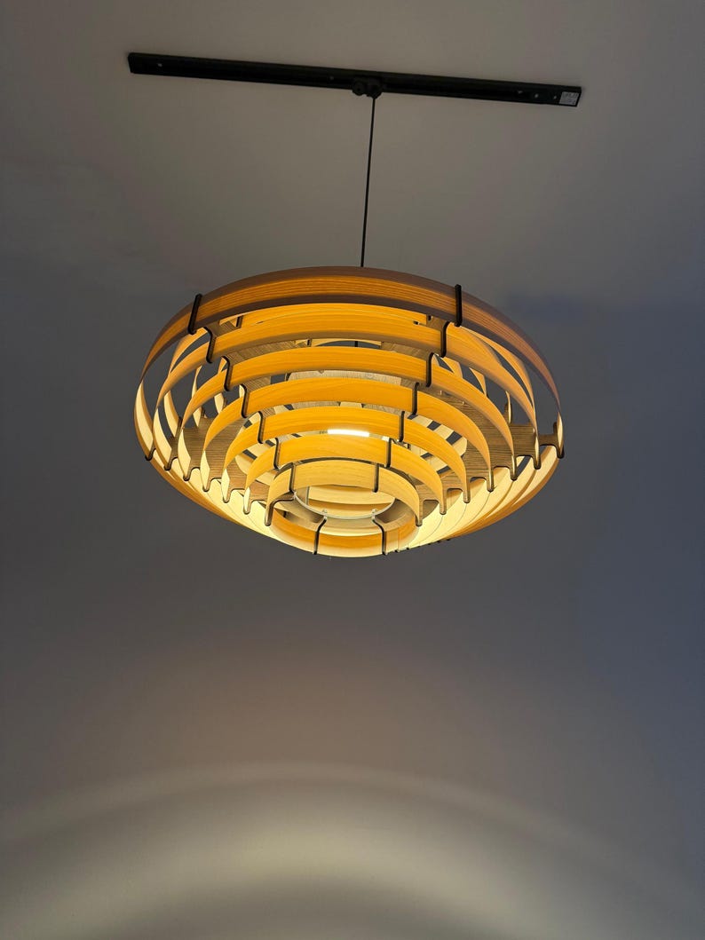 May include: A modern, wooden pendant light fixture. The light has a layered, circular design with warm, yellow light emanating from within. The fixture is suspended from a black cord and mounted on a black ceiling fixture.