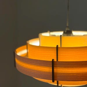Puede incluir: Una l&aacute;mpara colgante circular en capas con un brillo amarillo c&aacute;lido. La luz tiene una serie de bandas horizontales, con soportes de color marr&oacute;n oscuro. La l&aacute;mpara est&aacute; suspendida de un solo cable.