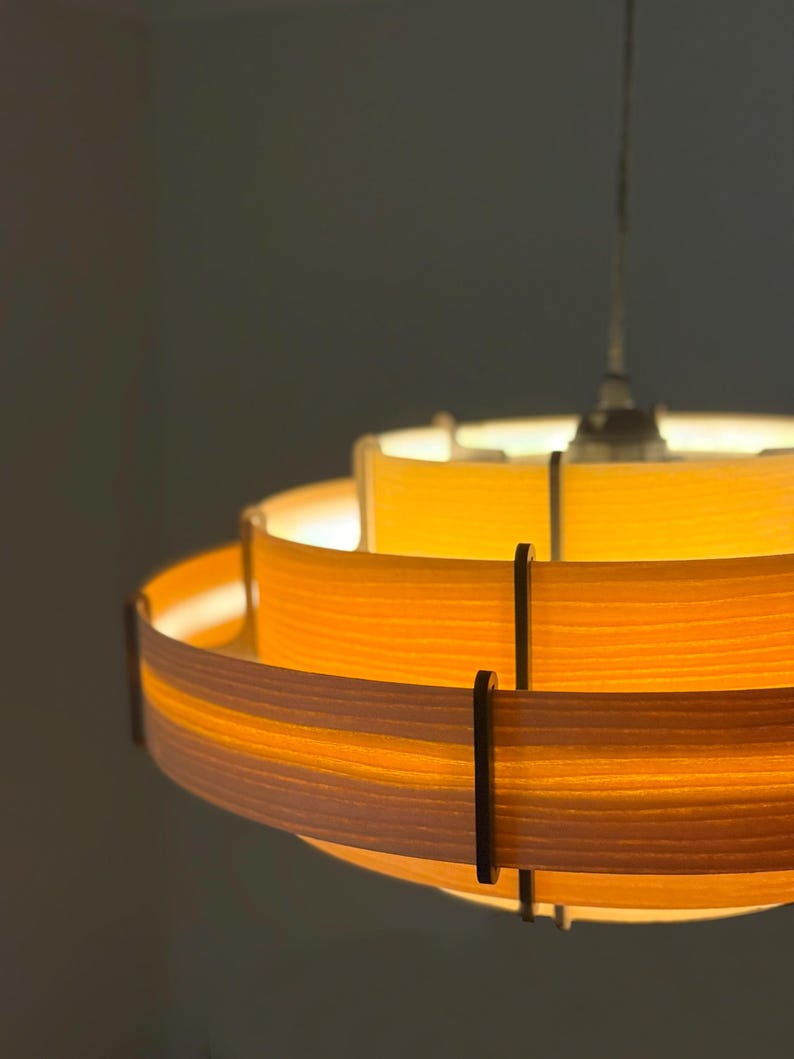 Puede incluir: Una l&aacute;mpara colgante circular en capas con un brillo amarillo c&aacute;lido. La luz tiene una serie de bandas horizontales, con soportes de color marr&oacute;n oscuro. La l&aacute;mpara est&aacute; suspendida de un solo cable.