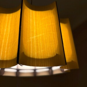 May include: A close-up of a hanging lampshade with a warm yellow glow. The lampshade has a layered design with a wooden texture, and black edges. The light source is visible at the bottom, creating a soft, ambient light.