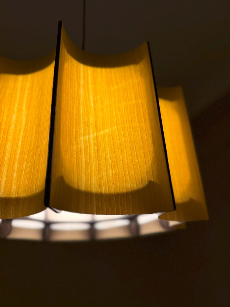 May include: A close-up of a hanging lampshade with a warm yellow glow. The lampshade has a layered design with a wooden texture, and black edges. The light source is visible at the bottom, creating a soft, ambient light.