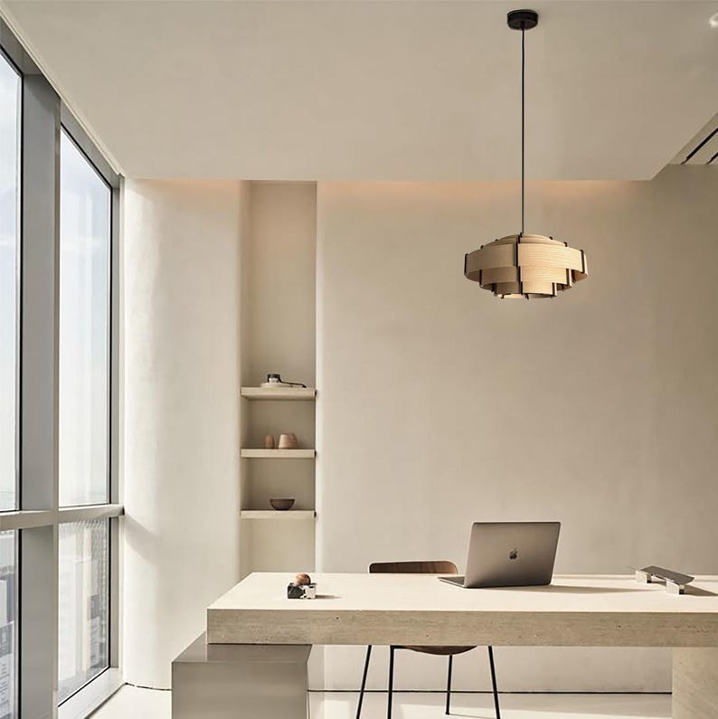 Puede incluir: Oficina moderna con l&aacute;mpara colgante de madera. Un escritorio con port&aacute;til y silla se encuentra cerca de una gran ventana. El espacio es minimalista, con tonos neutros y peque&ntilde;as estanter&iacute;as en la pared.
