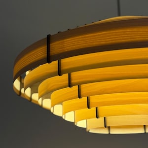 May include: A modern, layered wooden pendant light fixture. The light has a circular design with multiple horizontal wooden slats, creating a warm, ambient glow. The wood has a natural, light-yellow color, and the fixture is suspended from a dark cord.