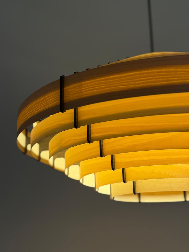 May include: A modern, layered wooden pendant light fixture. The light has a circular design with multiple horizontal wooden slats, creating a warm, ambient glow. The wood has a natural, light-yellow color, and the fixture is suspended from a dark cord.