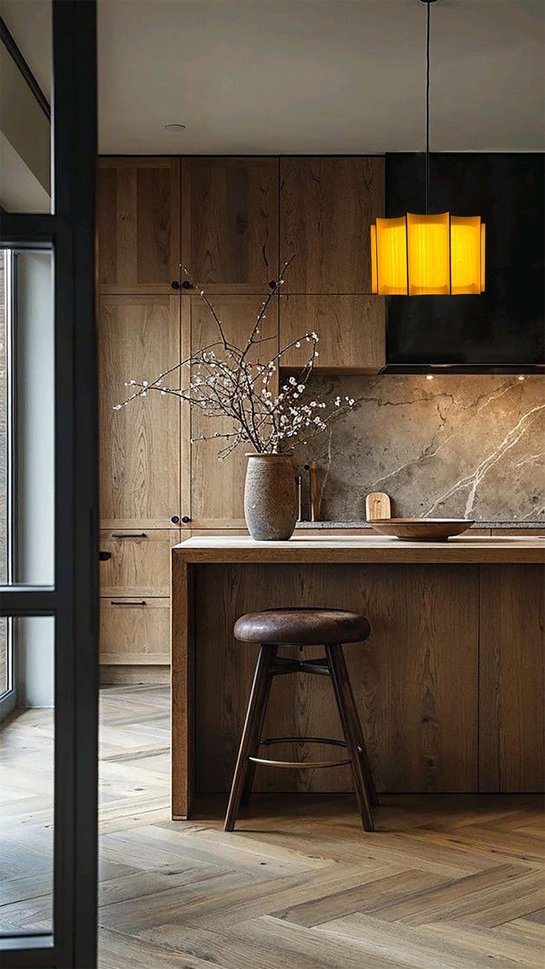 May include: A modern kitchen with wooden cabinets, a countertop, and a bar stool. A vase with branches sits on the countertop. A yellow rectangular pendant light hangs above the island. The floor has a herringbone pattern.