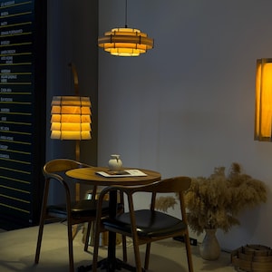 Puede incluir: Un comedor moderno con l&aacute;mparas colgantes de madera, una mesa redonda y dos sillas. Las luces tienen un dise&ntilde;o en capas, proyectando un brillo c&aacute;lido. Un jarr&oacute;n con plantas secas y una pieza decorativa de madera completan la escena.