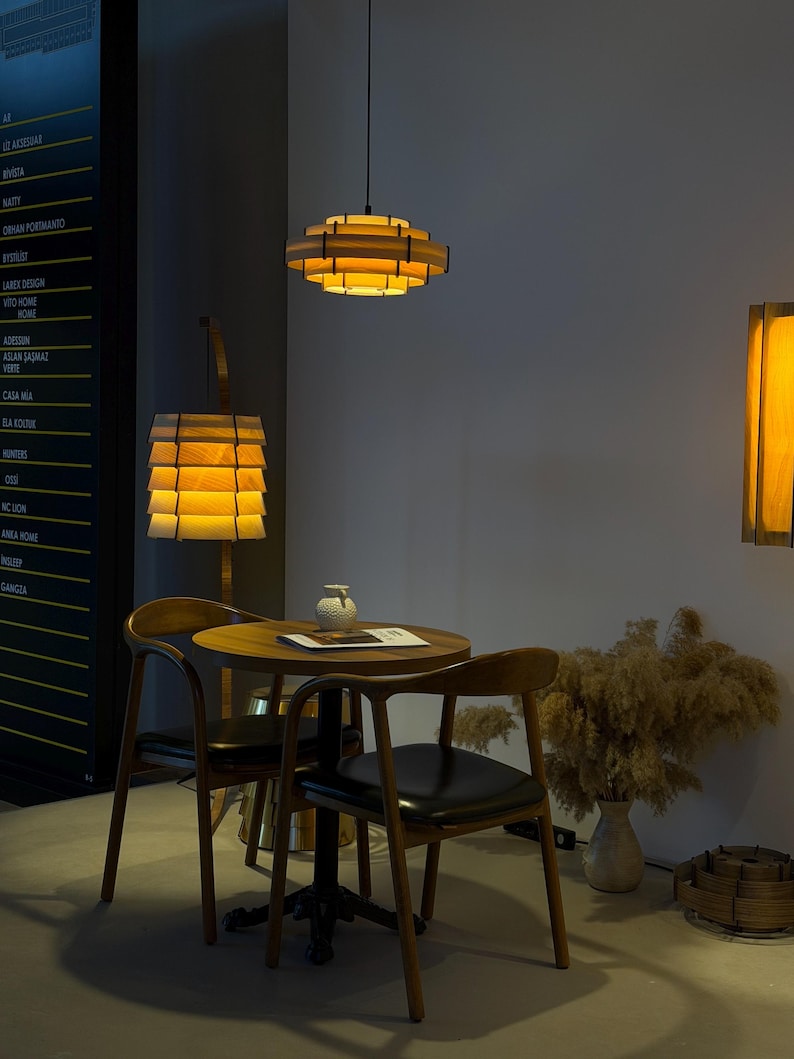 Puede incluir: Un comedor moderno con l&aacute;mparas colgantes de madera, una mesa redonda y dos sillas. Las luces tienen un dise&ntilde;o en capas, proyectando un brillo c&aacute;lido. Un jarr&oacute;n con plantas secas y una pieza decorativa de madera completan la escena.