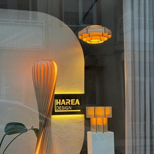 Puede incluir: Escaparate con l&aacute;mparas y luminarias modernas de madera. Se muestra una l&aacute;mpara de pie alta y retorcida, una l&aacute;mpara colgante circular y una peque&ntilde;a l&aacute;mpara de mesa. El letrero en el escaparate dice "HAREA DESIGN". Las l&aacute;mparas emiten una luz c&aacute;lida y acogedora.