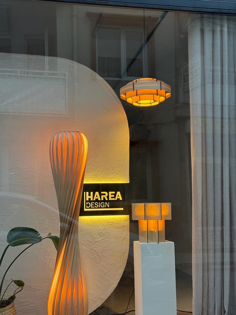 Puede incluir: Escaparate con l&aacute;mparas y luminarias modernas de madera. Se muestra una l&aacute;mpara de pie alta y retorcida, una l&aacute;mpara colgante circular y una peque&ntilde;a l&aacute;mpara de mesa. El letrero en el escaparate dice "HAREA DESIGN". Las l&aacute;mparas emiten una luz c&aacute;lida y acogedora.