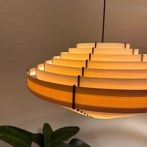 May include: A modern, layered wooden pendant light fixture. The light has a circular, tiered design with warm, amber-toned illumination. The fixture is suspended from a dark cord, and the design is visible against a neutral background.