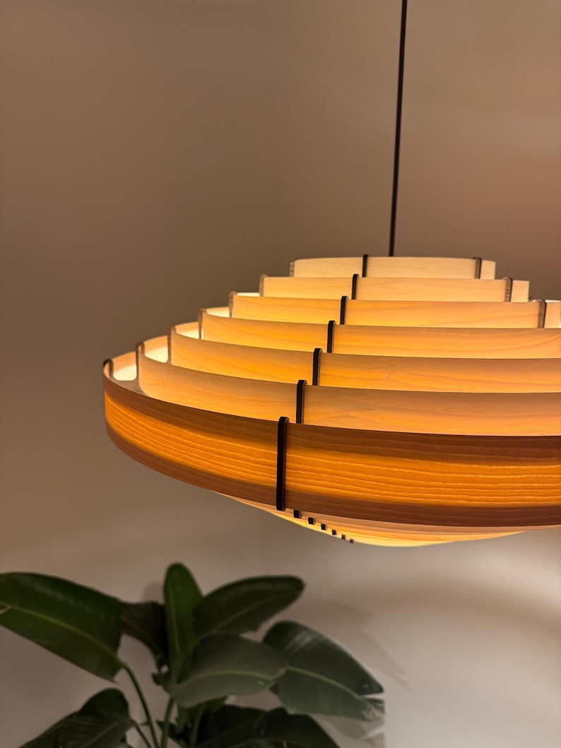 May include: A modern, layered wooden pendant light fixture. The light has a circular, tiered design with warm, amber-toned illumination. The fixture is suspended from a dark cord, and the design is visible against a neutral background.