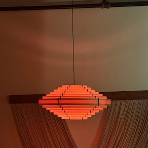 May include: A modern, geometric pendant light fixture. The light is made of layered wooden slats in shades of orange and red, suspended from a black cord. The fixture is diamond-shaped and emits a warm glow, suitable for interior lighting.