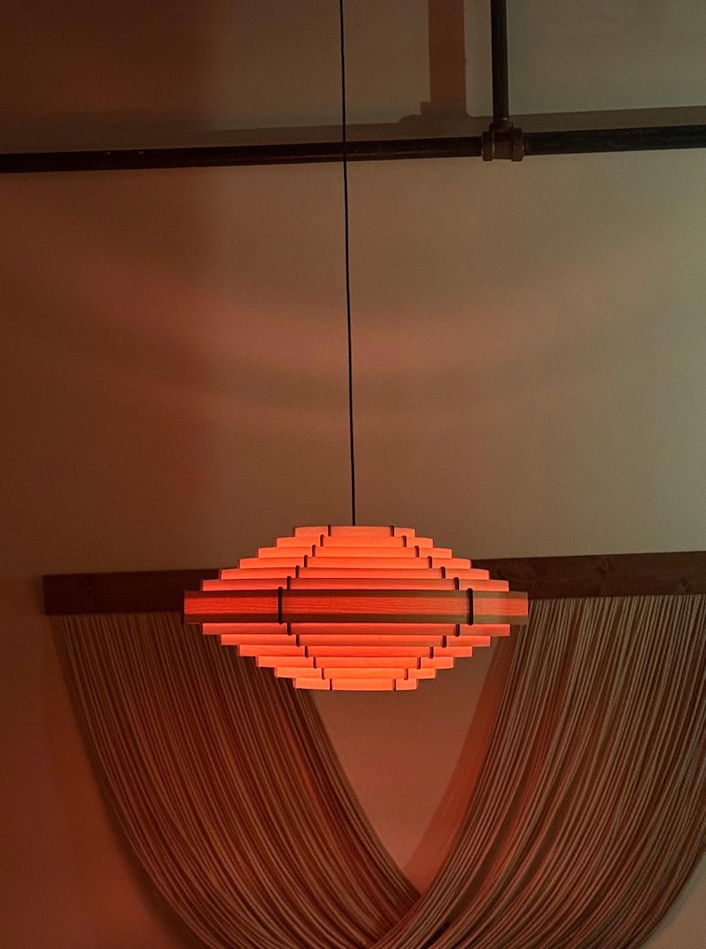 May include: A modern, geometric pendant light fixture. The light is made of layered wooden slats in shades of orange and red, suspended from a black cord. The fixture is diamond-shaped and emits a warm glow, suitable for interior lighting.