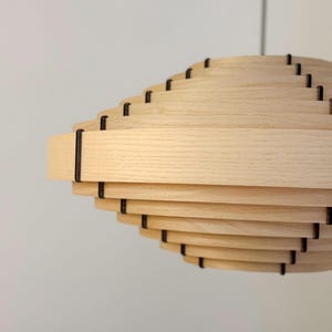 May include: A light-colored wooden pendant light fixture with a layered, spiral design. The fixture is made of horizontal wooden slats held together by dark connectors. The light hangs from a thin, dark cord against a white background.