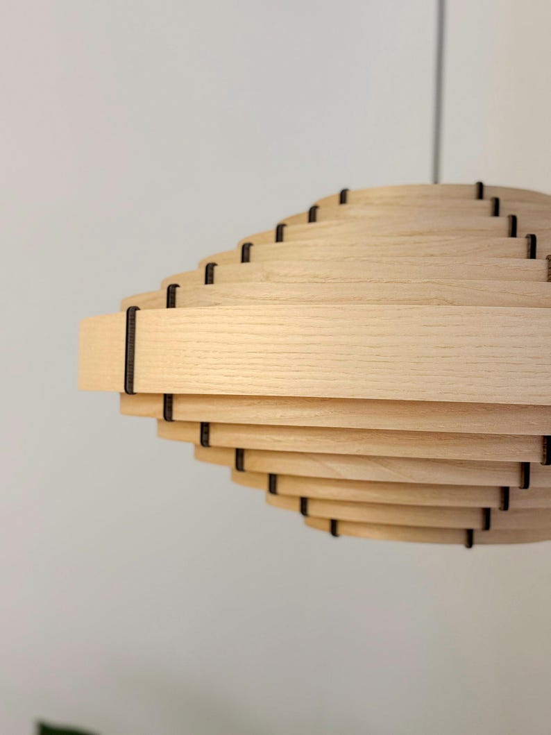 May include: A light-colored wooden pendant light fixture with a layered, spiral design. The fixture is made of horizontal wooden slats held together by dark connectors. The light hangs from a thin, dark cord against a white background.