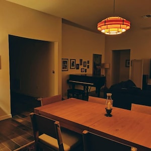 Puede incluir: Un comedor con una gran mesa de madera y sillas. Una l&aacute;mpara colgante roja y naranja cuelga sobre la mesa. Un piano y cuadros enmarcados son visibles en el fondo. Una linterna de velas est&aacute; sobre la mesa.