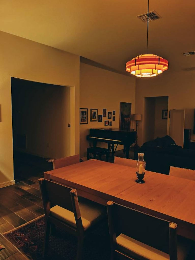 Puede incluir: Un comedor con una gran mesa de madera y sillas. Una l&aacute;mpara colgante roja y naranja cuelga sobre la mesa. Un piano y cuadros enmarcados son visibles en el fondo. Una linterna de velas est&aacute; sobre la mesa.