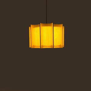 May include: A modern pendant light fixture with a rectangular shade. The shade is made of vertical wooden slats with a warm yellow glow emanating from within. The fixture hangs from a thin black cord against a dark brown background.