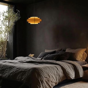 Puede incluir: Un dormitorio con una est&eacute;tica oscura y melanc&oacute;lica. Una cama en capas con ropa de cama gris es el punto focal. Una l&aacute;mpara colgante geom&eacute;trica y en capas cuelga sobre la cama. Se ve una ventana con plantas de bamb&uacute;.