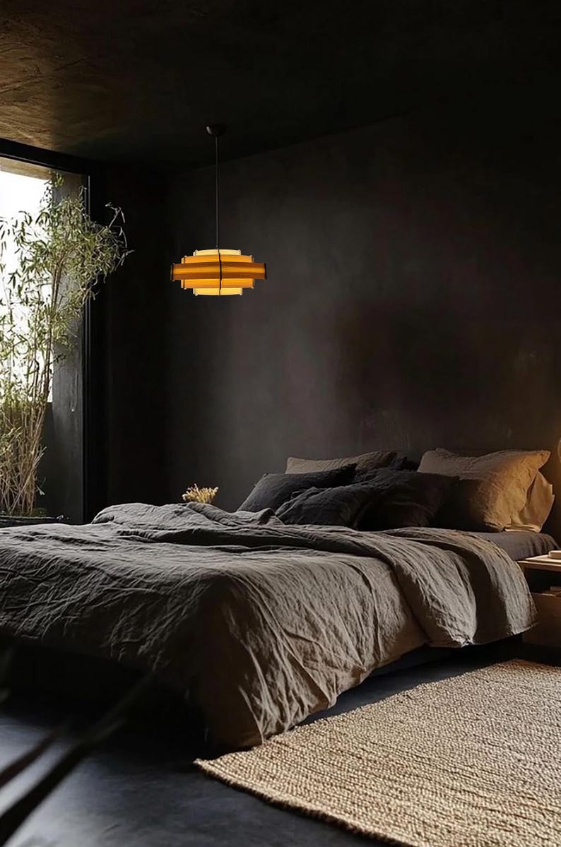 Puede incluir: Un dormitorio con una est&eacute;tica oscura y melanc&oacute;lica. Una cama en capas con ropa de cama gris es el punto focal. Una l&aacute;mpara colgante geom&eacute;trica y en capas cuelga sobre la cama. Se ve una ventana con plantas de bamb&uacute;.