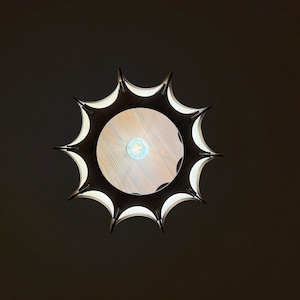 May include: Overhead view of a modern, sunburst-shaped light fixture. The fixture has a dark, spiky outer frame with white interior accents. The center features a light-colored wooden surface with a visible lightbulb.